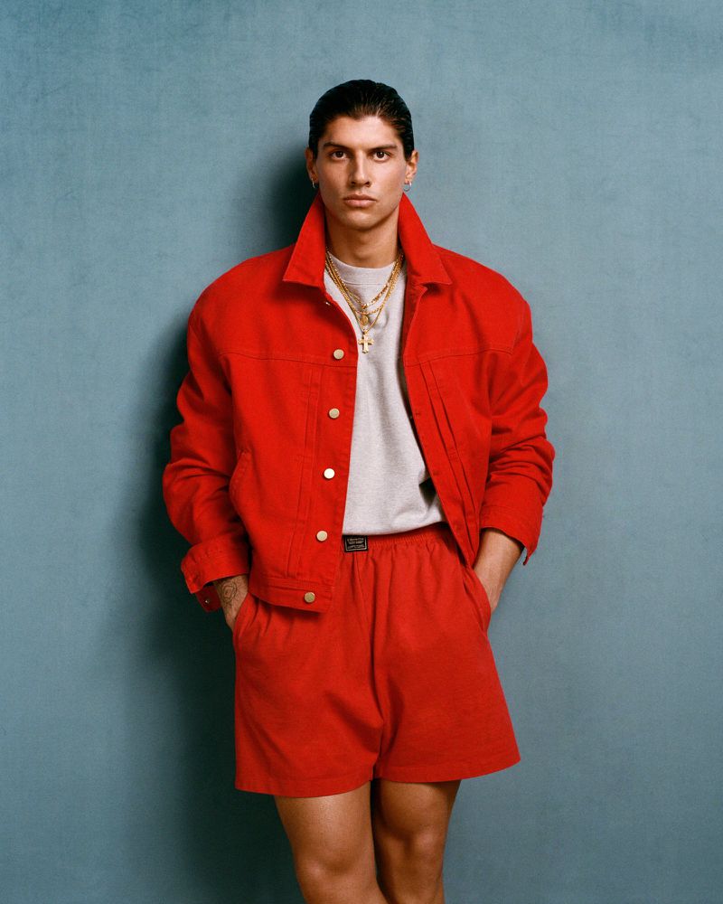 Portrait of a young adult man in a red denim jacket and shorts, paired with gold chains, against a textured blue backdrop.