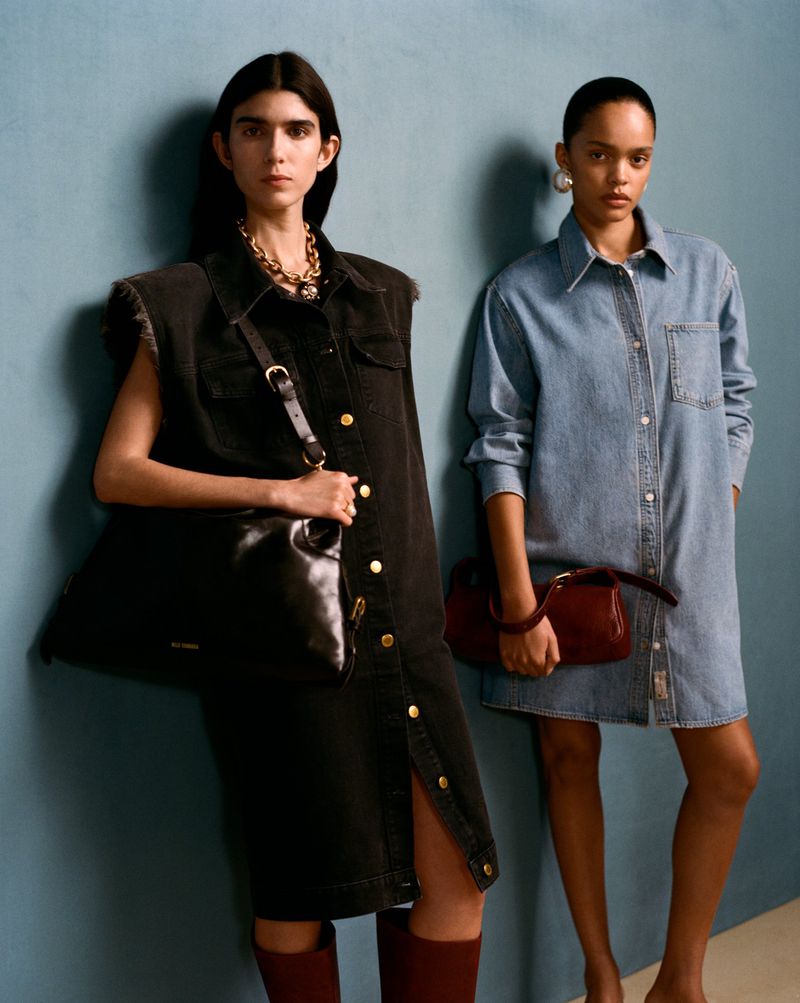Two women in denim shirt dresses pose against a blue wall, one carrying a black leather handbag, the other a red leather handbag.