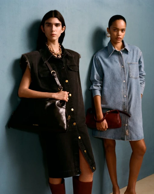 Two women in denim shirt dresses pose against a blue wall, one carrying a black leather handbag, the other a red leather handbag.