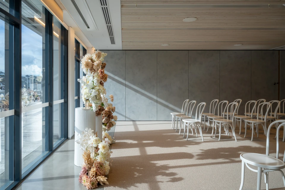 Indoor event space setup with rows of white chairs, large windows, and a floral arrangement on a white column for a ceremony.