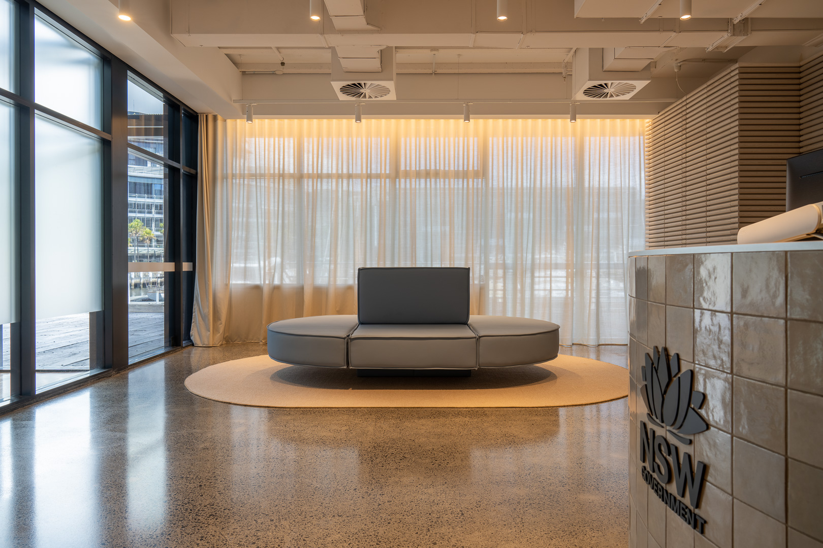Modern office lobby featuring a round couch on a rug, large windows, and a reception desk with the NSW Government logo.