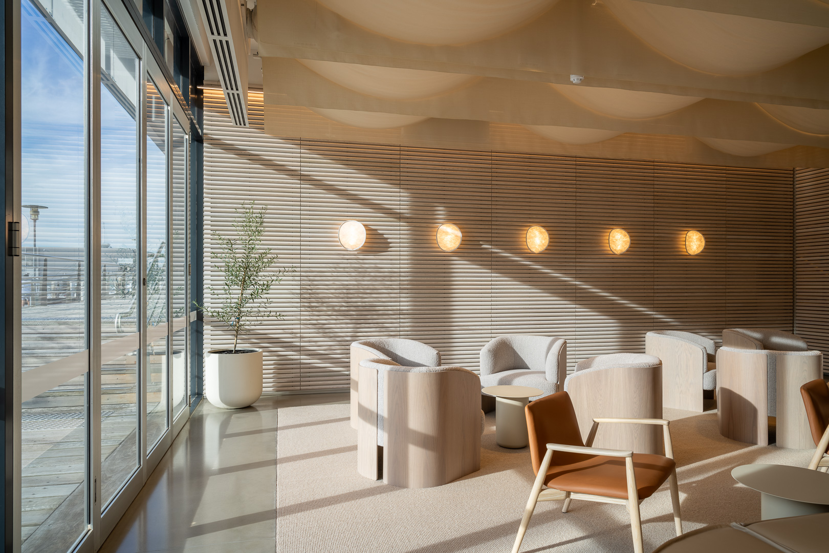 Indoor lounge area featuring modern chairs, tables, and lighting with floor-to-ceiling windows and a potted plant near the windows.