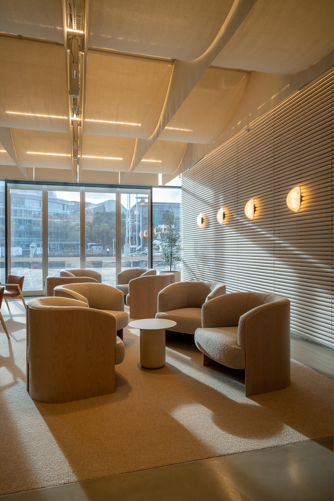 Beige armchairs arranged around a round table in a modern lounge area with a textured wall and a large window.