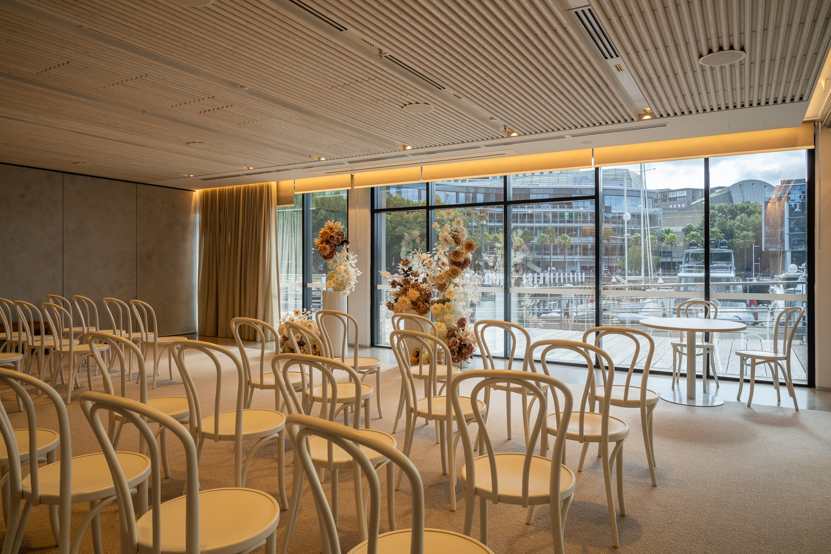 Indoor event space with rows of chairs, floral arrangements, and large windows overlooking a harbor, creating an elegant setting.