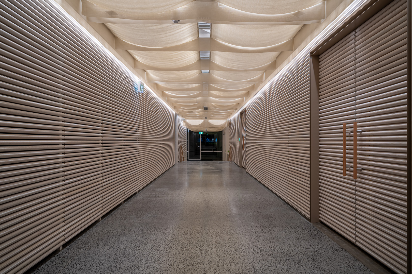 Indoor shot of a long hallway with light grey walls with parallel lines, a patterned ceiling, and a light grey speckled floor.