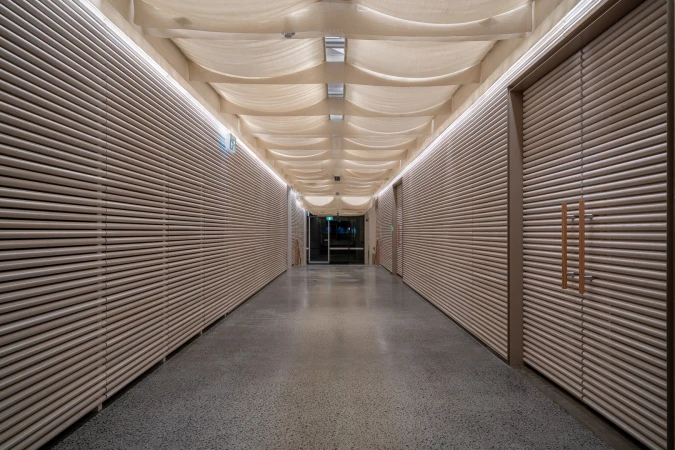 Indoor shot of a long hallway with light grey walls with parallel lines, a patterned ceiling, and a light grey speckled floor.