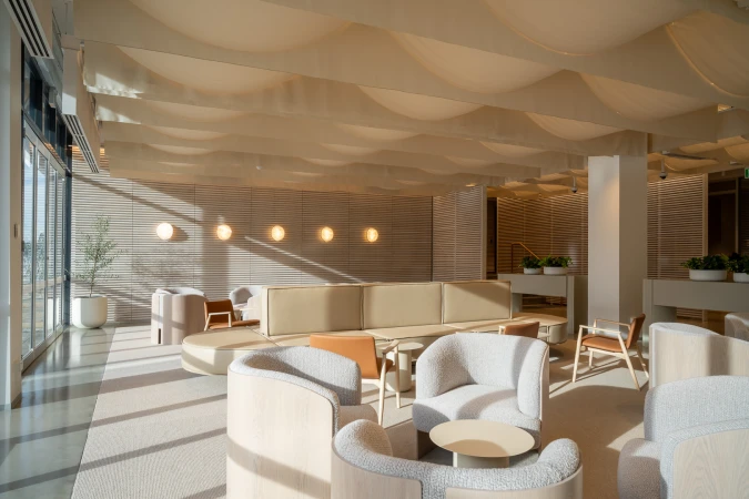 Modern interior waiting area with curved armchairs, low tables, slatted wood walls, and a fabric-draped ceiling.