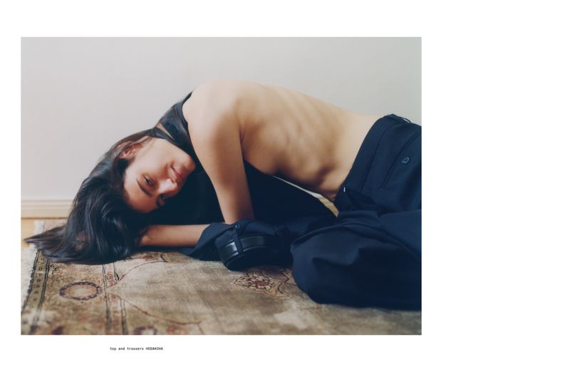 Brunette woman lies on an ornate rug wearing a dark top and loose, dark trousers in a minimalist setting with neutral colors and soft light.