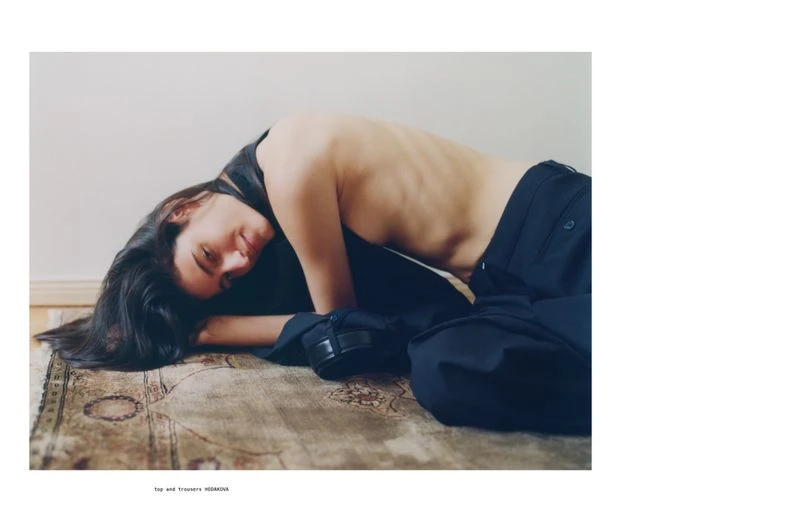 Brunette woman lies on an ornate rug wearing a dark top and loose, dark trousers in a minimalist setting with neutral colors and soft light.