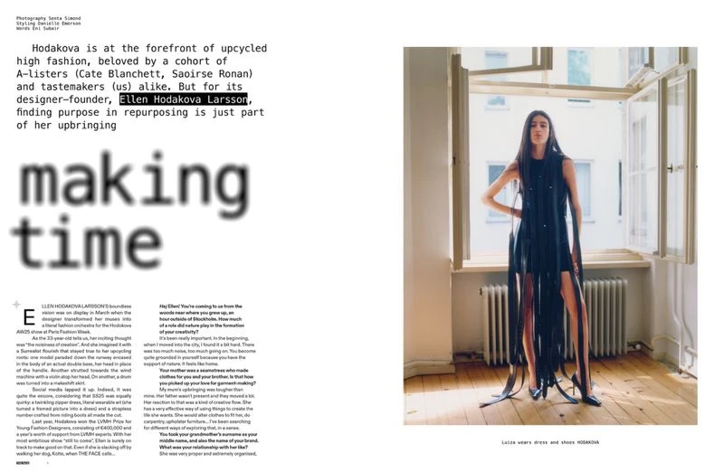 Editorial fashion shot of a woman in a black dress with fringe standing near an open window in a minimally decorated room.
