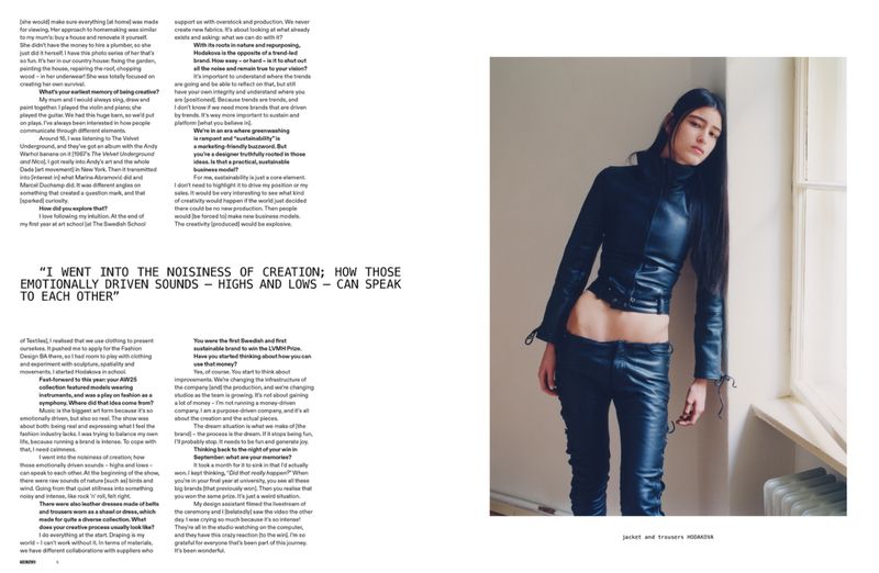 A slender young woman in a black leather outfit leans against a wall by a window in a full shot fashion editorial-style portrait.