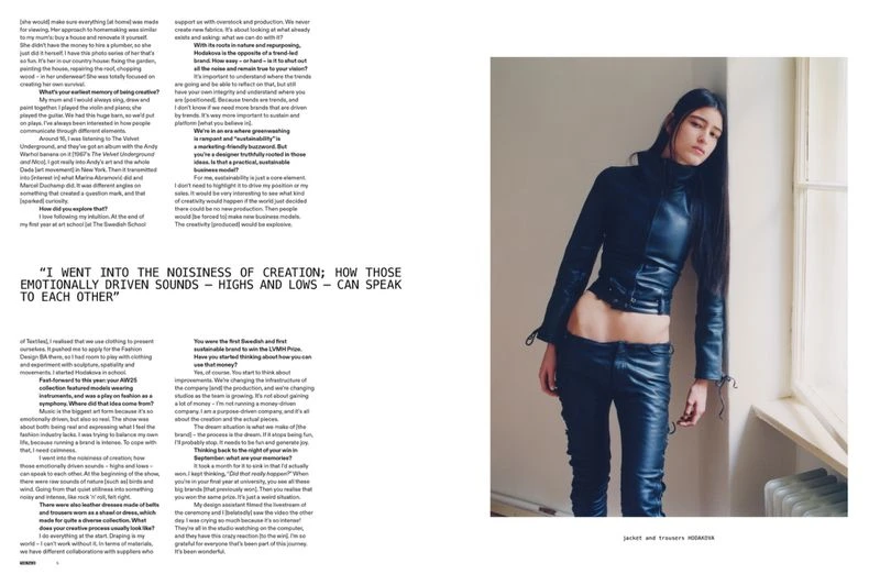 A slender young woman in a black leather outfit leans against a wall by a window in a full shot fashion editorial-style portrait.