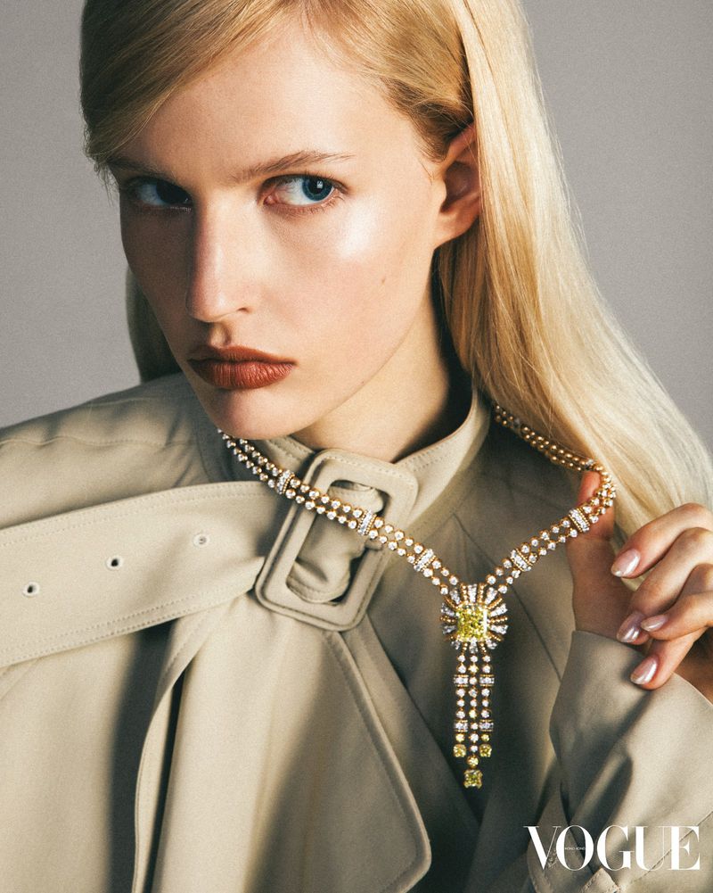 Elegant woman with blonde hair and light skin wearing a beige trench coat and a yellow diamond necklace while posing indoors.