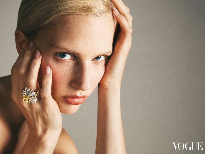 Close-up of a blonde woman with her hands near her face, wearing a large jeweled ring; studio shot with grey background and soft lighting.