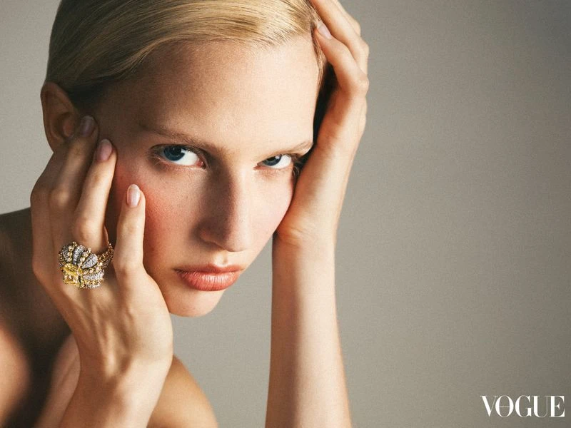 Close-up of a blonde woman with her hands near her face, wearing a large jeweled ring; studio shot with grey background and soft lighting.