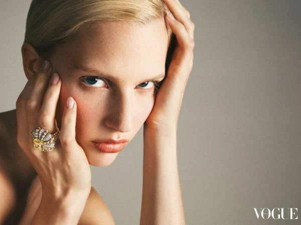 Close-up of a blonde woman with her hands near her face, wearing a large jeweled ring; studio shot with grey background and soft lighting.
