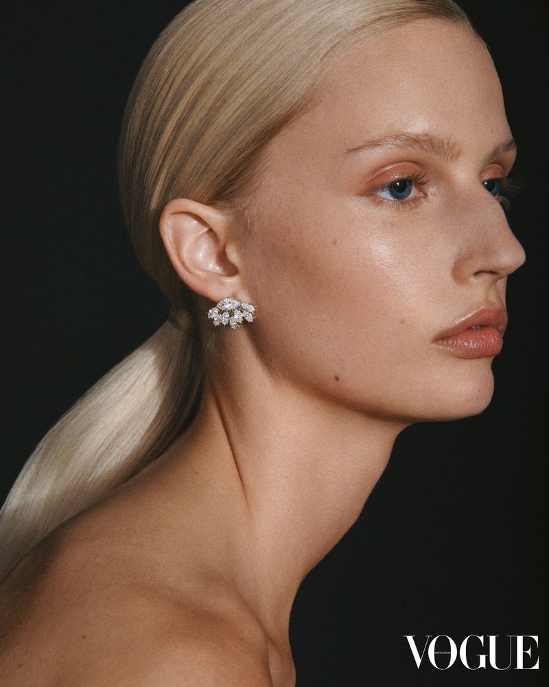 Close-up beauty shot of a blonde woman with fair skin, wearing elegant floral earrings, posing in a dark studio environment, Vogue logo.