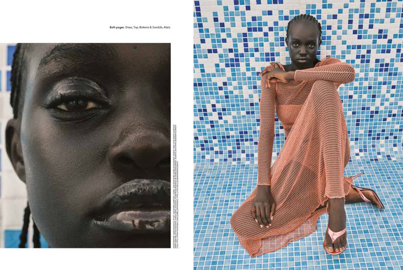 Fashion shot of a black woman in a peach-colored, net dress and sandals, seated in front of a blue mosaic tile wall with neutral expression.
