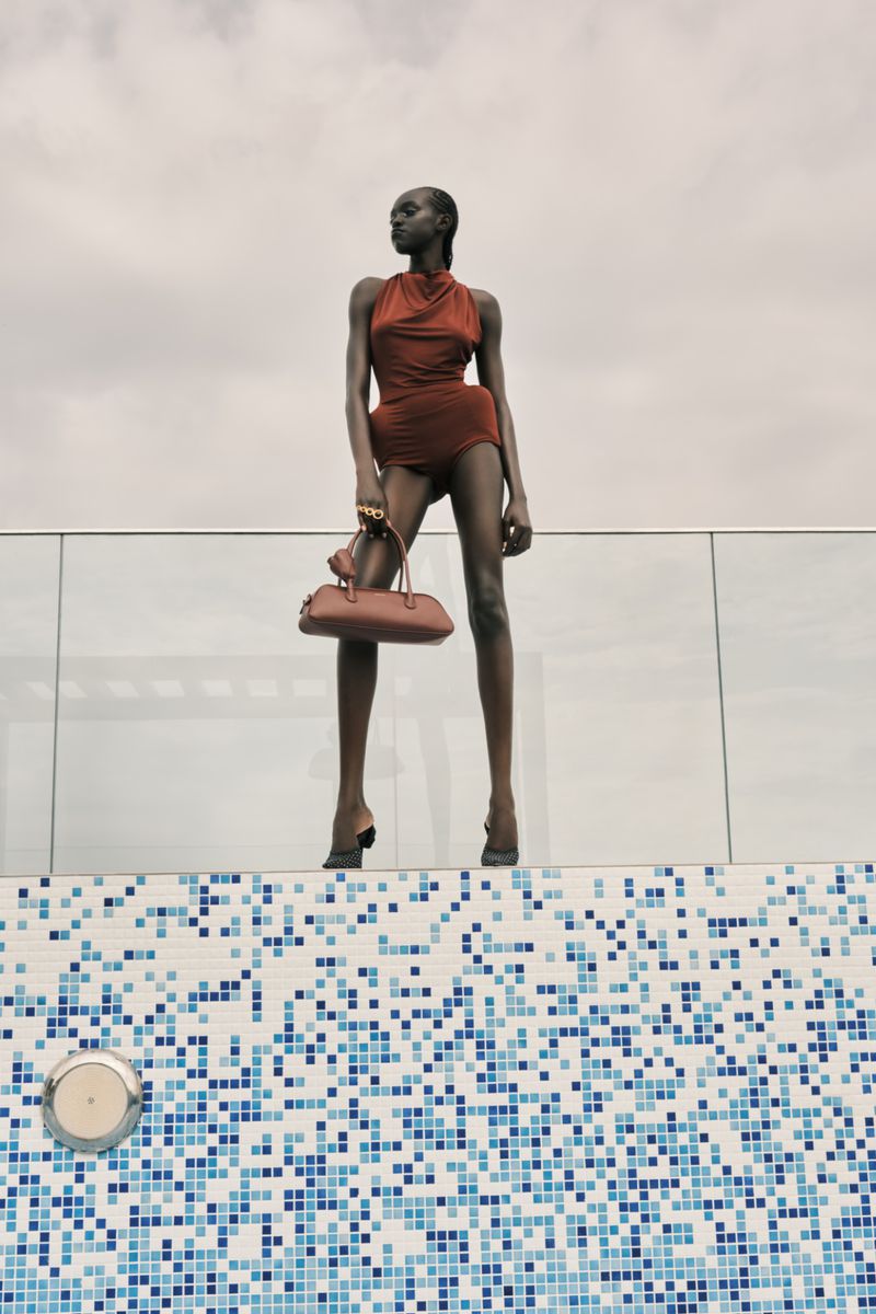 An adult female fashion model poses in a copper-colored halter romper, heels, and carrying a purse against a blue mosaic pool background.