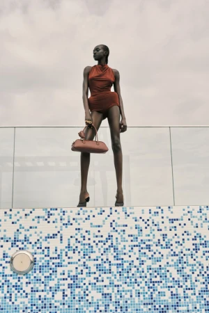 An adult female fashion model poses in a copper-colored halter romper, heels, and carrying a purse against a blue mosaic pool background.