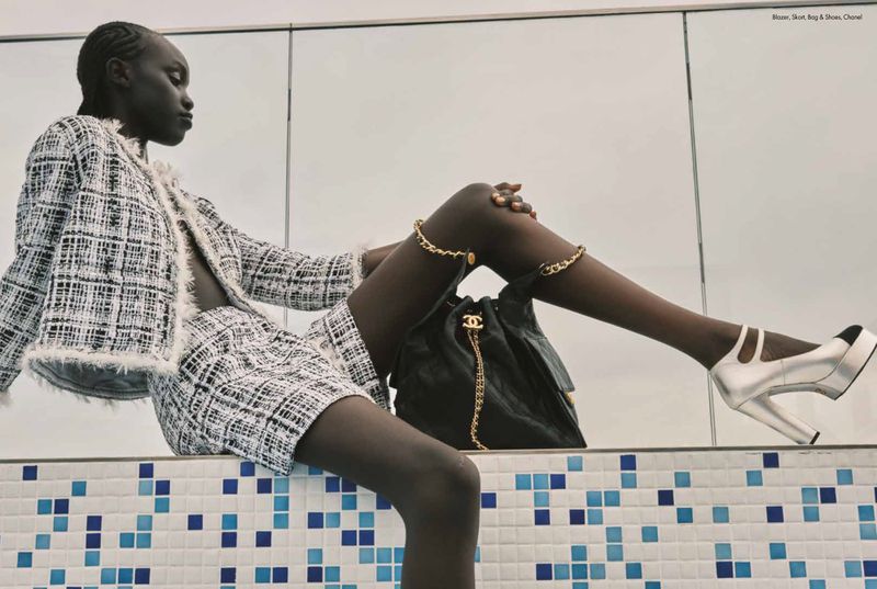 Fashion editorial shot featuring a Black woman wearing a tweed skirt suit with a Chanel bag and ankle bracelets in a studio setting.