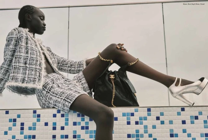 Fashion editorial shot featuring a Black woman wearing a tweed skirt suit with a Chanel bag and ankle bracelets in a studio setting.