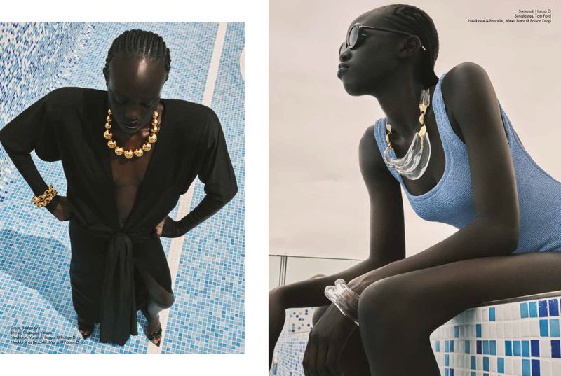 Two dark-skinned women pose in swimwear and statement gold jewelry near a blue-tiled pool, showcasing fashion and elegance in a bright setting.