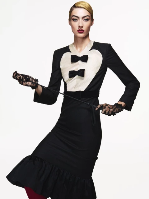 An adult female model with golden blonde hair posing in a black dress with black lace gloves, holding a riding crop, against a white background.