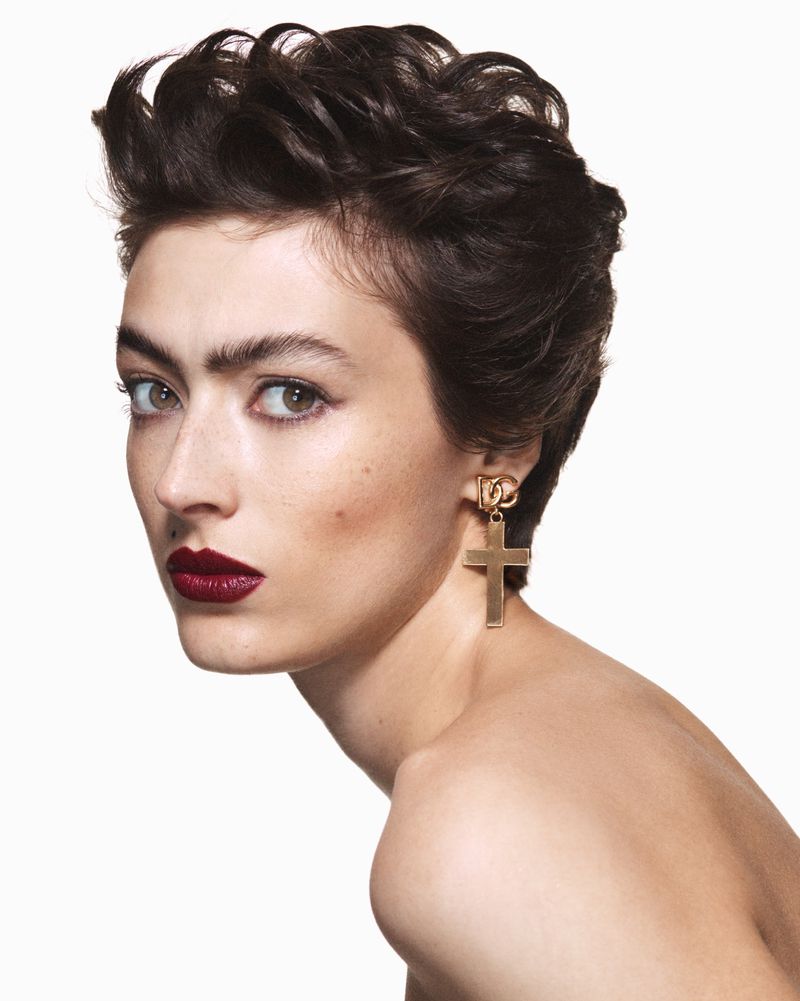 Portrait of a woman with dark hair and dark lipstick wearing a gold cross earring against a white background in a beauty shot.