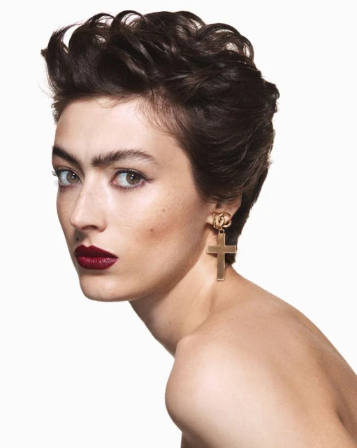 Portrait of a woman with dark hair and dark lipstick wearing a gold cross earring against a white background in a beauty shot.