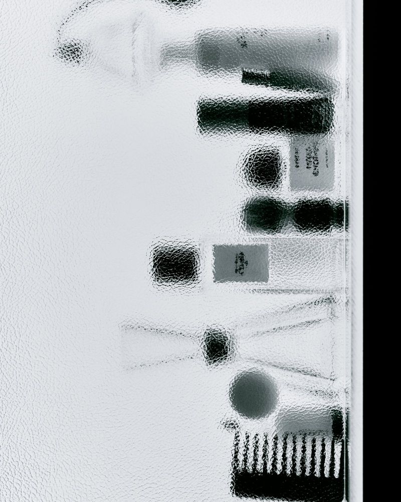 Overhead shot of skincare and cosmetic products behind textured glass, featuring tubes, containers, and a makeup brush.