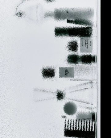 Overhead shot of skincare and cosmetic products behind textured glass, featuring tubes, containers, and a makeup brush.
