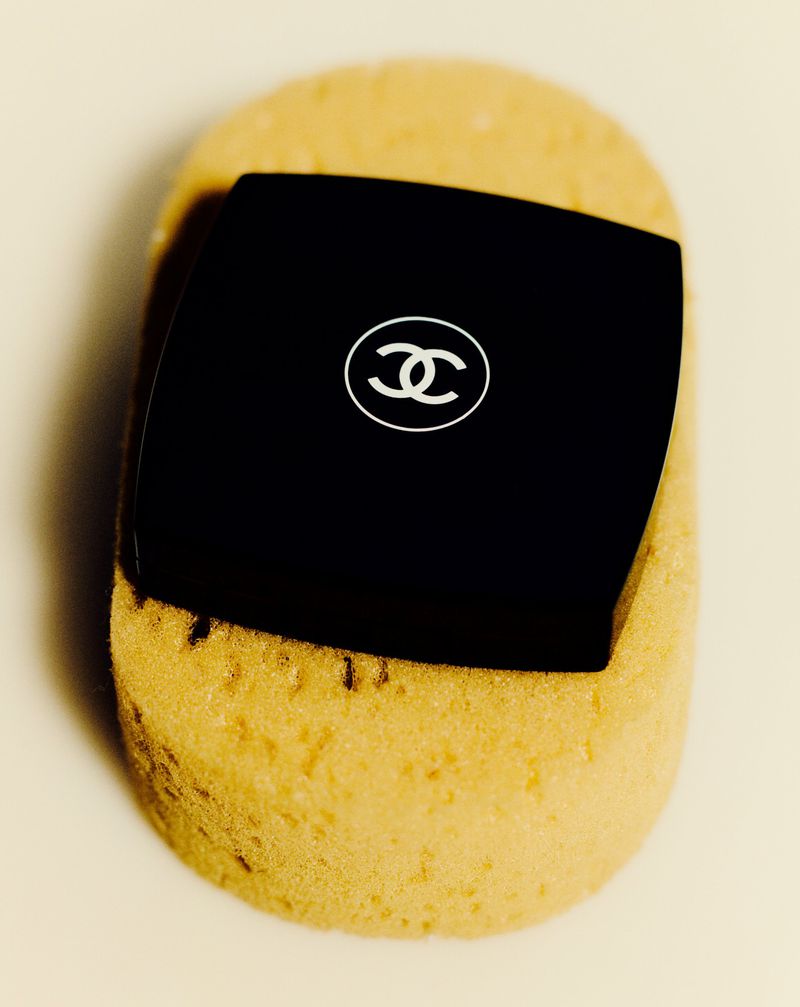 High-angle shot of a black Chanel compact on a textured tan sponge atop a white surface, showcasing luxury beauty product.