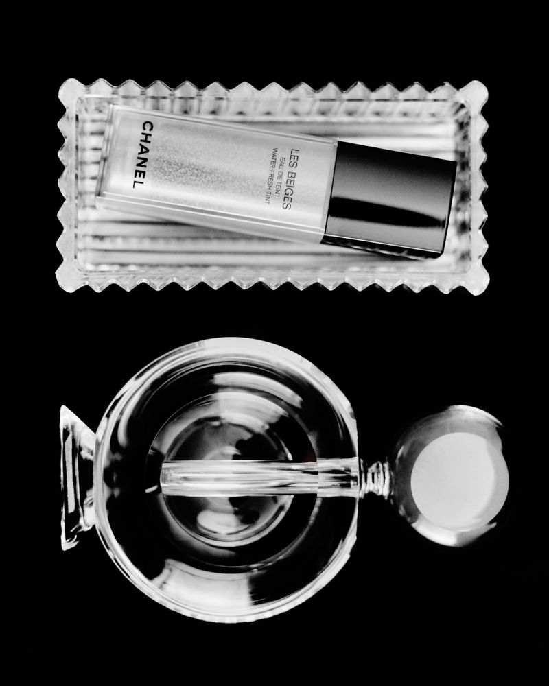 Overhead studio shot of Chanel foundation on a glass tray, along with a vintage ice cream dish against a solid black backdrop.