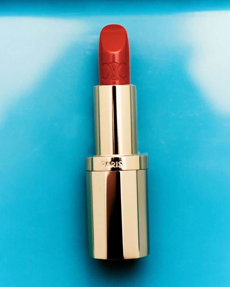 Close-up of a red and gold lipstick tube on a blue background. The tube is centered, with a blue and white backdrop.