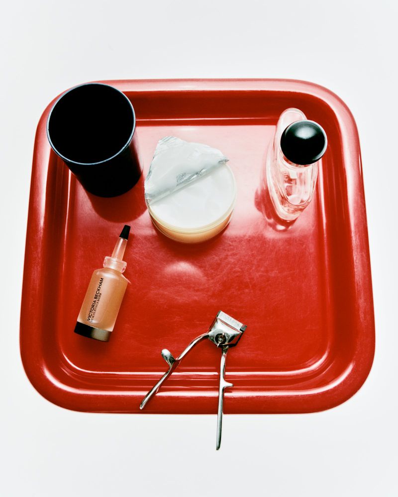 Overhead shot of beauty products on a red tray including lotion, an eyelash curler, and a black cup, all on a white surface.