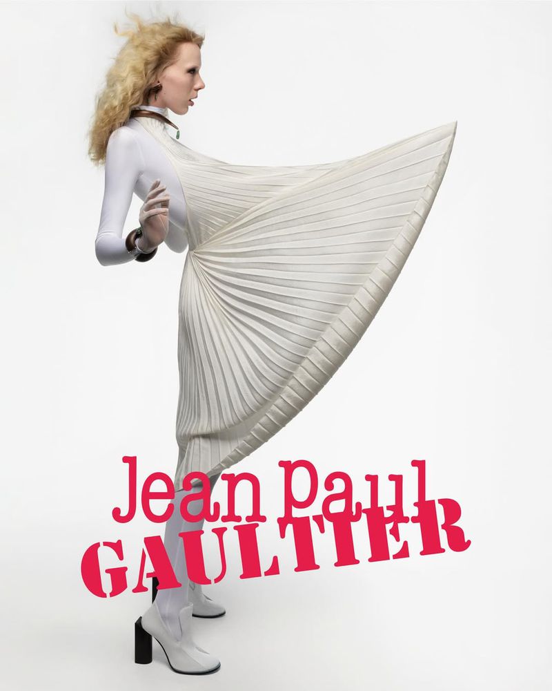 Blonde woman poses in profile wearing a white pleated dress and white heeled boots, against a plain white backdrop with brand text.