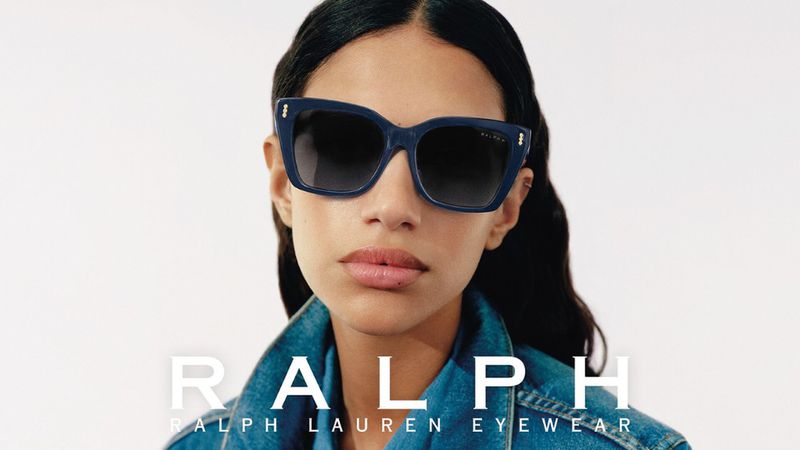 Close-up of an adult woman wearing blue-framed sunglasses, a denim jacket, and dark hair, against a white background with brand logo text.