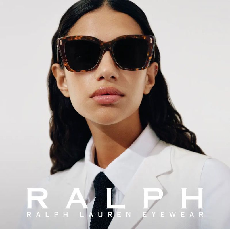 A brunette adult female poses in front of a bright, seamless backdrop, wearing tortoise shell sunglasses, white blazer, and a dark tie.