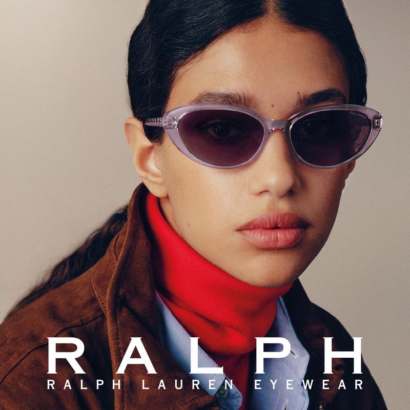 Model wearing translucent sunglasses with a red turtleneck and brown jacket against a neutral backdrop