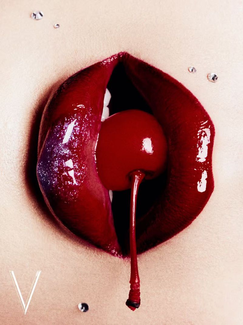 Close-up of red lips holding a cherry in the mouth, with small rhinestones around the mouth area. Bright, sensual beauty image.