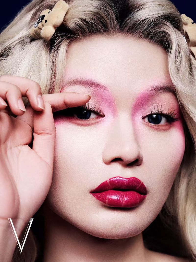 Close-up portrait of an adult female model with dramatic pink eye makeup and glossy lips, styled with plush bear hair accessories.
