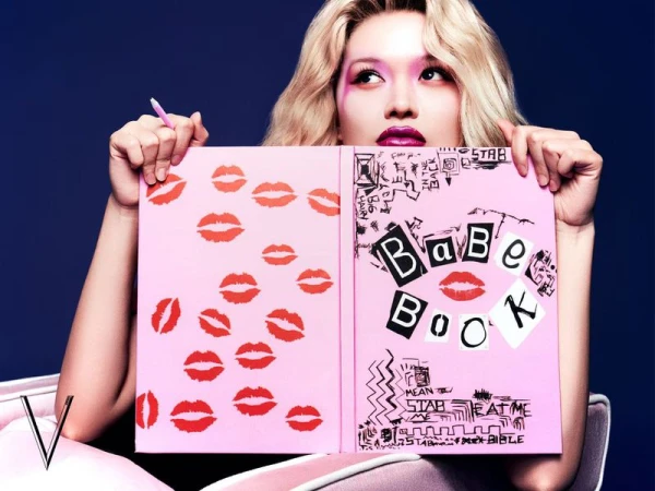 Woman with blond hair holding a pink book with red lips and text on it in front of a blue background in a medium studio shot.