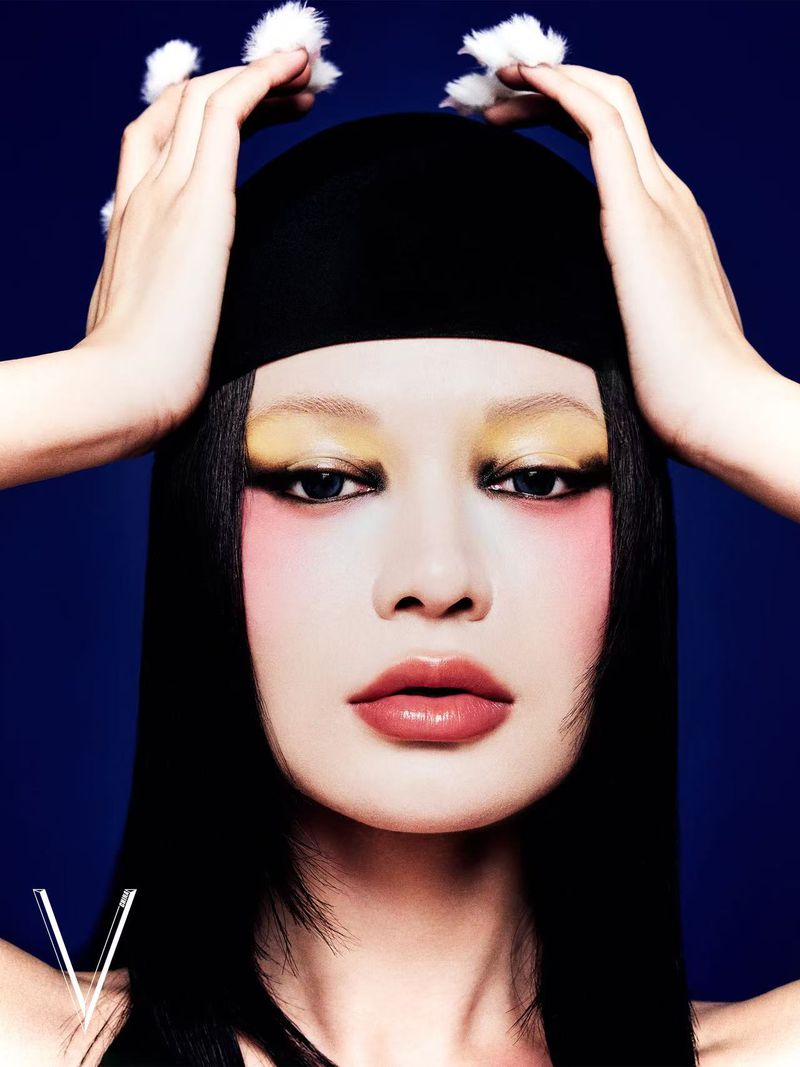 Close-up portrait of an adult female model with dramatic makeup, a black head covering, and fuzzy white pompoms against a blue background.