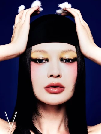 Close-up portrait of an adult female model with dramatic makeup, a black head covering, and fuzzy white pompoms against a blue background.