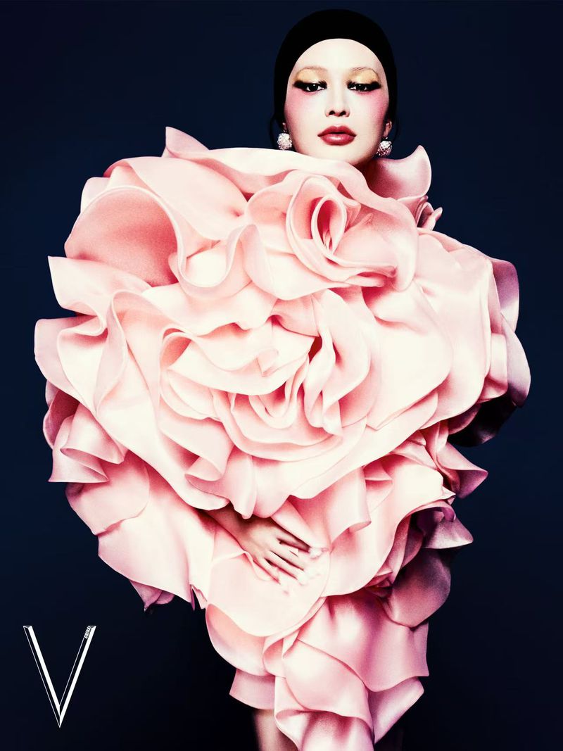 Fashion editorial shot of woman in a large pink ruffled dress and black head covering against a dark blue background, with 'V' logo.