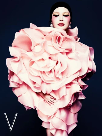 Fashion editorial shot of woman in a large pink ruffled dress and black head covering against a dark blue background, with 'V' logo.