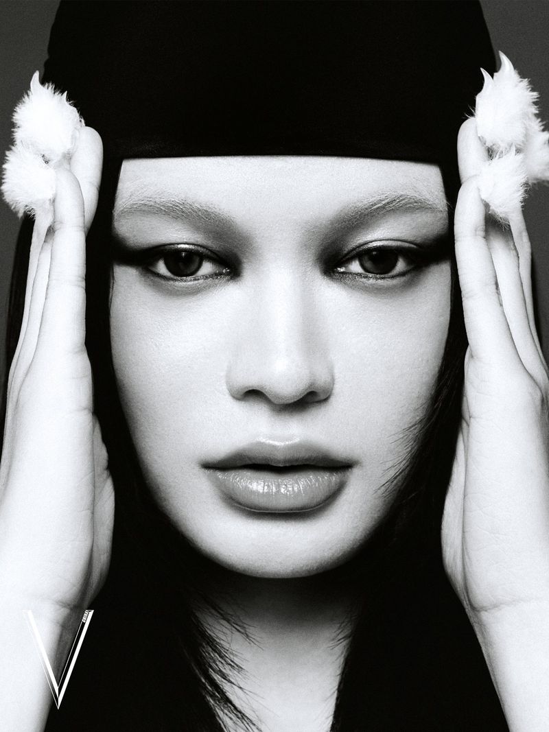 Close-up portrait of a woman with hands framing her face, wearing a black head covering adorned with white fur accents.