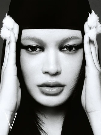 Close-up portrait of a woman with hands framing her face, wearing a black head covering adorned with white fur accents.