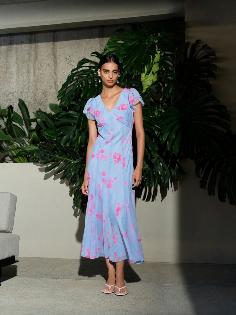 A fair-skinned young woman stands in front of large tropical leaves, modeling a light blue floral dress and pale pink flip-flops.